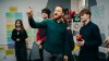 A man in a green shirt gestures while speaking to a small group of young people in a classroom, with colourful notes and posters on the walls.